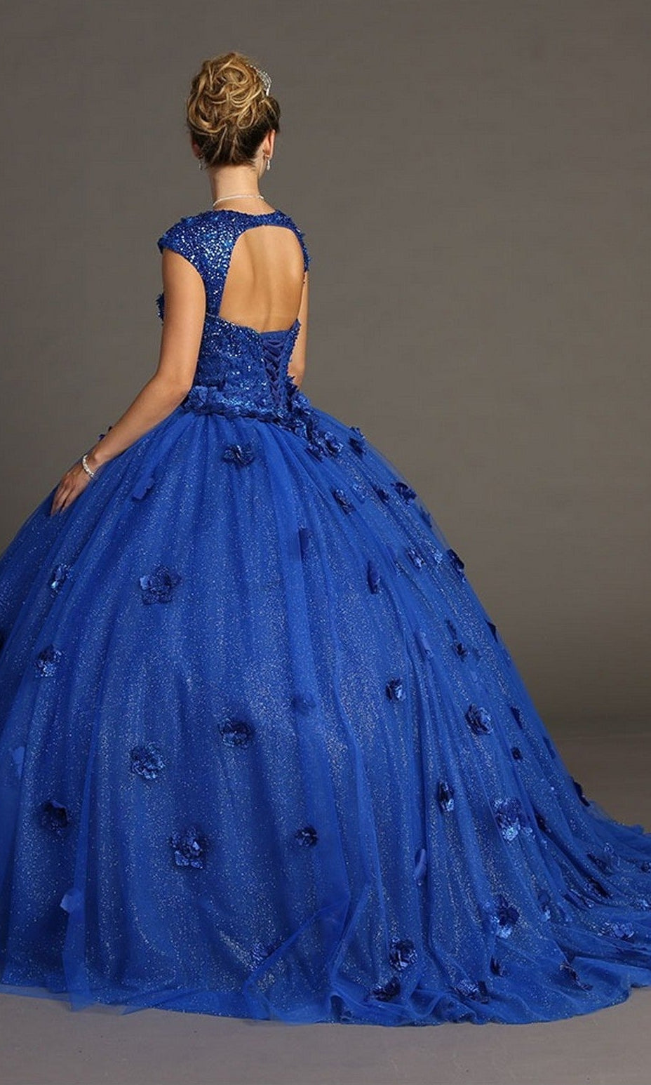 A woman wearing the Quinceanera Dress CHF3068 By Chicas—a sparkling royal blue gown with 3D floral accents and a low back—stands against a plain gray background.